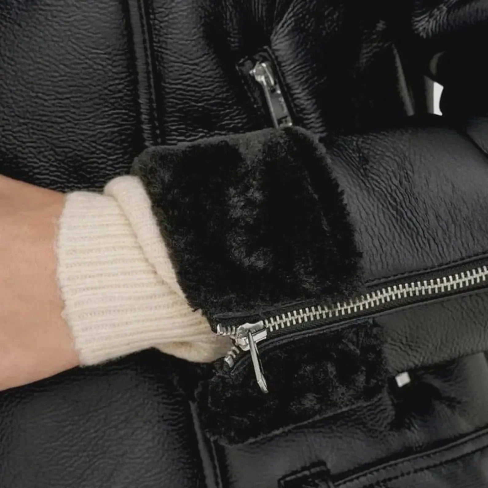 Close-up of a person wearing a black leather jacket with fur-lined cuffs and a beige knit sleeve.